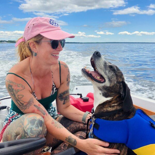 Adoption Process 3 A woman wearing a pink cap and sunglasses smiles at her dog, who is wearing a blue life jacket and sitting beside her in a boat. The background shows a calm body of water and a partly cloudy sky. Both the woman and the dog look happy and relaxed.