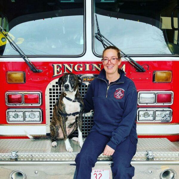 Adoption Process 1 A firefighter in uniform sits next to a dog on the front bumper of a red fire truck, with "East Providence" and "Engine 1" written on it. They both look at the camera, and the firefighter is smiling. The fire truck has various tools and equipment visible in the background.