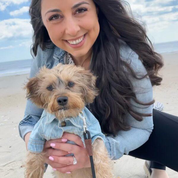 Adoption Process 2 A woman with long dark hair smiles while holding a small dog. Both the woman and the dog are wearing denim jackets. They are at a beach with the ocean and a cloudy sky in the background.