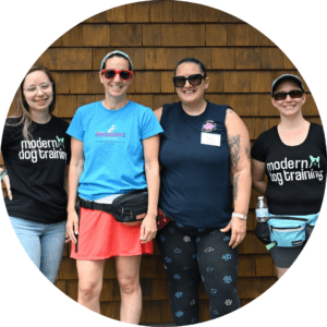 Our Partners 8 Four people stand in front of a wooden wall, smiling. Two on the ends wear "modern dog training" shirts, while the two in the middle wear different shirts. Some have sunglasses on, and one person is carrying a waist pouch and a water bottle.