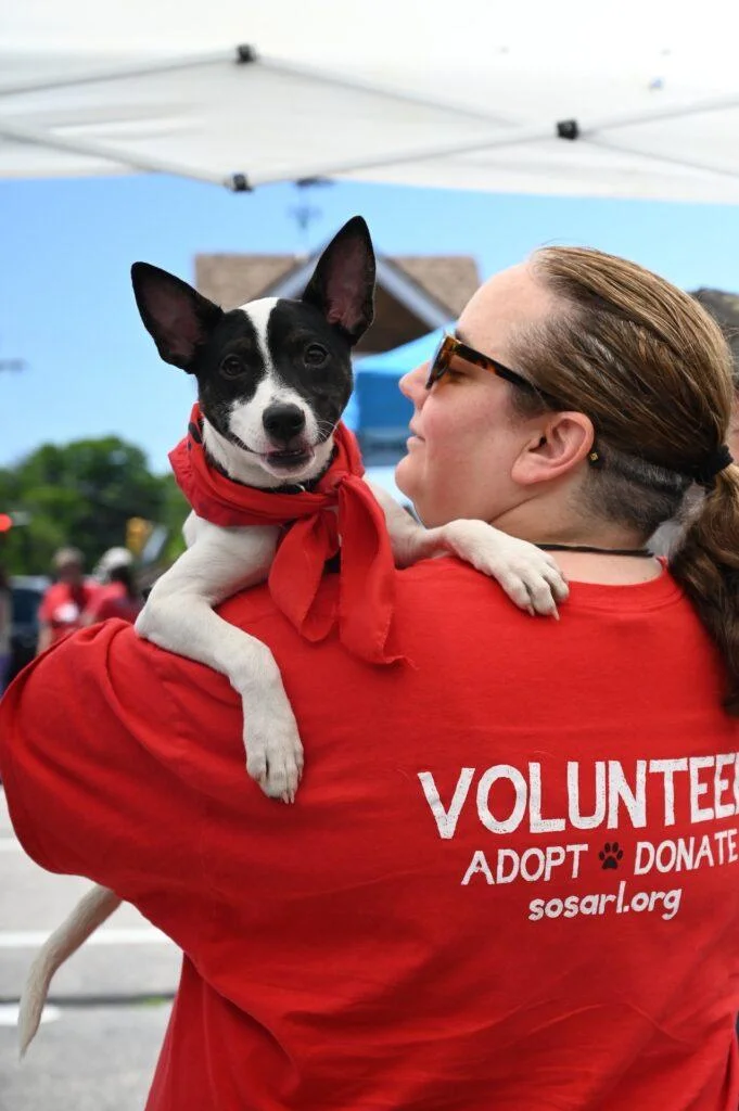A person wearing a red shirt with white text that reads "VOLUNTEER ADOPT DONATE sosarl.org" is holding a small, black and white dog. The dog is wearing a red bandana around its neck. They appear to be at an outdoor event under a tent.