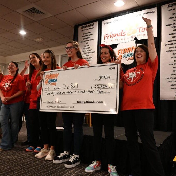A group of people wearing red T-shirts stands on a stage holding a large ceremonial check. The check is made out to "One Soul" for $20,304.84, dated 3/23/24, from Funny 4 Funds. A white banner in the background displays sponsors' logos and information.