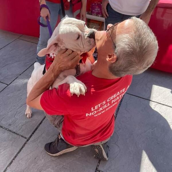 thank-you-volunteers A man wearing a red shirt and glasses is kneeling and embracing a white dog that is licking his face. Other people are in the background. The text on the man's shirt reads "Let's create a no-kill nation.